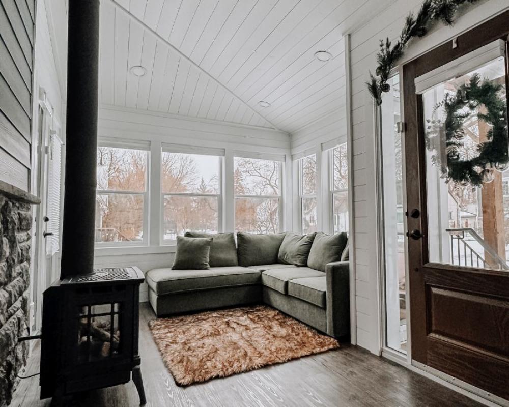 Cozy enclosed 3 season sun room featuring green sectional, stone accent wall, and wood stove